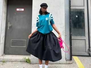 Woman wears black Adidas Samba sneakers, full black skirt, Adidas soccer jersey and a page-boy cap.