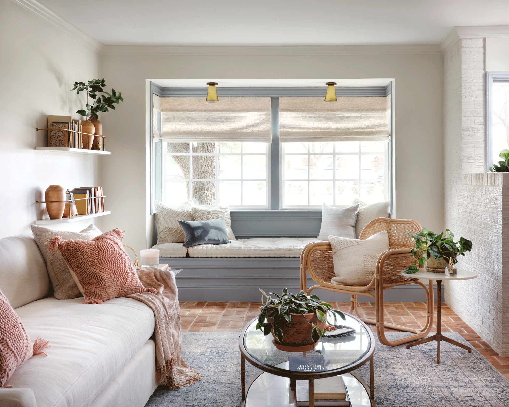 A peach, white and grey living room scheme with shelves, grey window seat, rattan chair and white linen sofa with pink cushions and small round coffee table