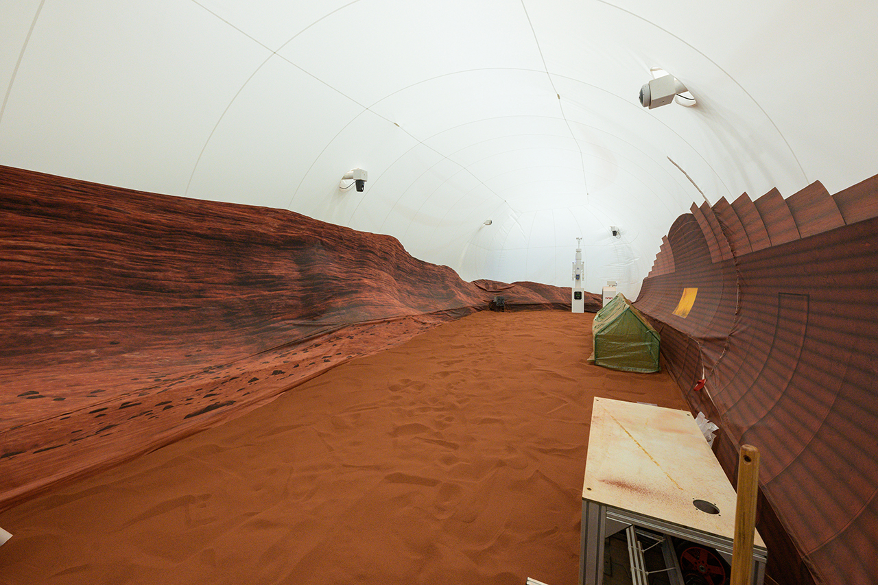 a long hallway with a red sandy flooring and printouts on the partitions to acquire it looks as if a martian landscape