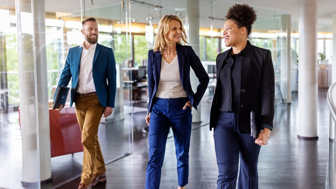 Three smiling employees walk side by side through an office. 