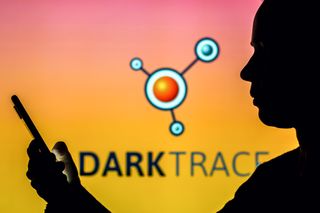 Darktrace logo in the background on an orange and pink gradient screen, with the silhouette of a man in the foreground holding a smartphone out in front of him