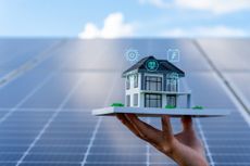 Home energy concept: Hand holds model house in front of solar panels.