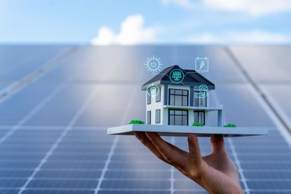 Home energy concept: Hand holds model house in front of solar panels.