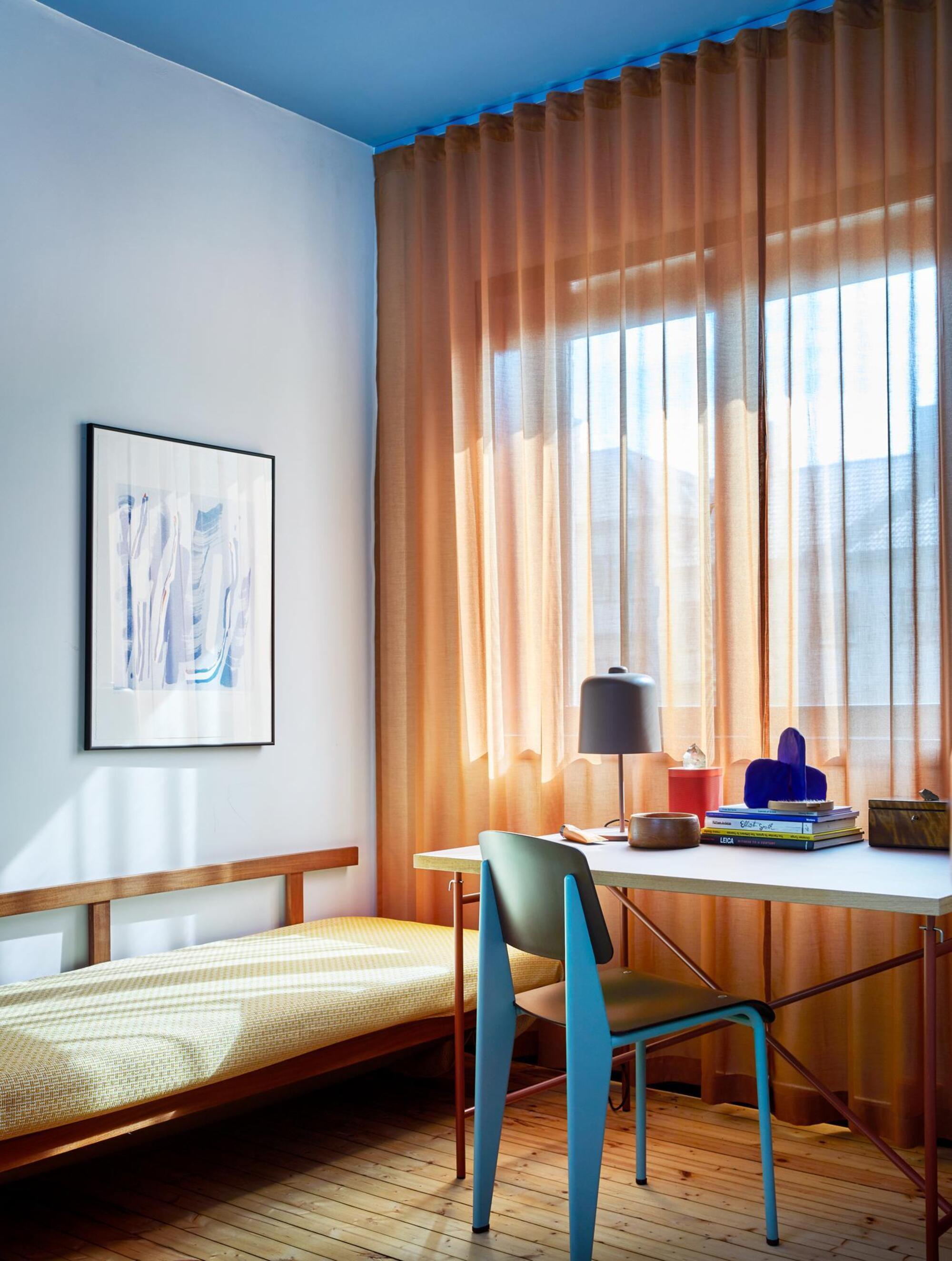 colorful bedroom with blue ceiling, light blue walls, sheer orange curtains, desk with blue chair and styled with books, lamp, and decor, and yellow single bed