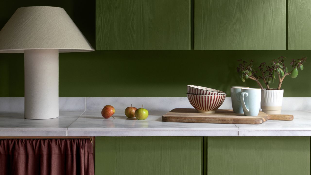 Close-up image of a kitchen counter. The cabinets are painted in a bright olive green and the countertops are made of a white marble. There is a large, fabric table lamp and ceramic bowls and mugs on the counter.
