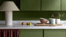 Close-up image of a kitchen counter. The cabinets are painted in a bright olive green and the countertops are made of a white marble. There is a large, fabric table lamp and ceramic bowls and mugs on the counter.