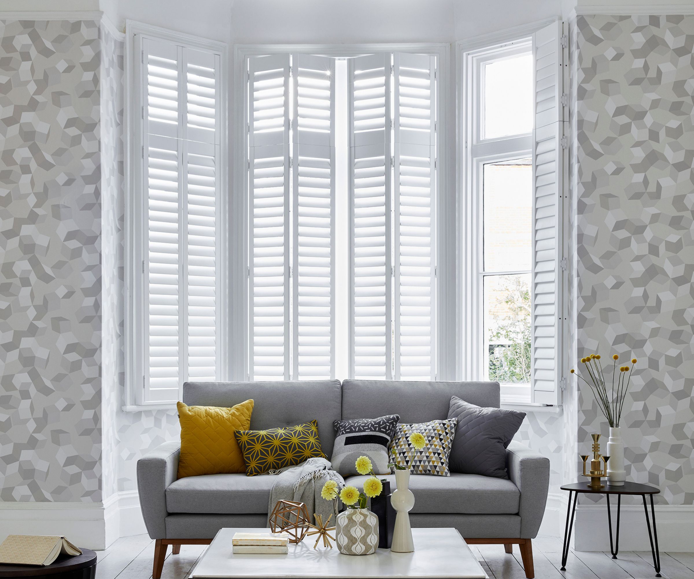 A bright living room with tall white bay windows fitted with plantation shutters. A grey sofa with patterned cushions sits against a wall with geometric wallpaper, styled with yellow and white vases.