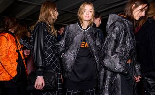 Four models, two wearing black shiny jackets, one wearing an orange jacket and one wearing a black and white check jacket with leather sleeves