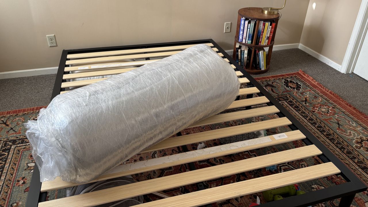 Queen-sized Casper The One Mattress rolled up and wrapped in plastic, positioned at an angle on our tester's slatted bed frame with an ornate rug underneath