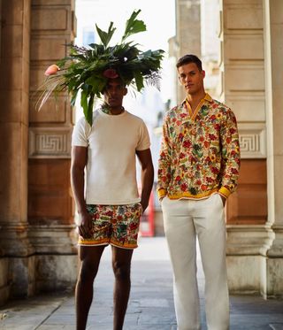 Two men wearing flowered patterned clothing and a large flower crown on a London street