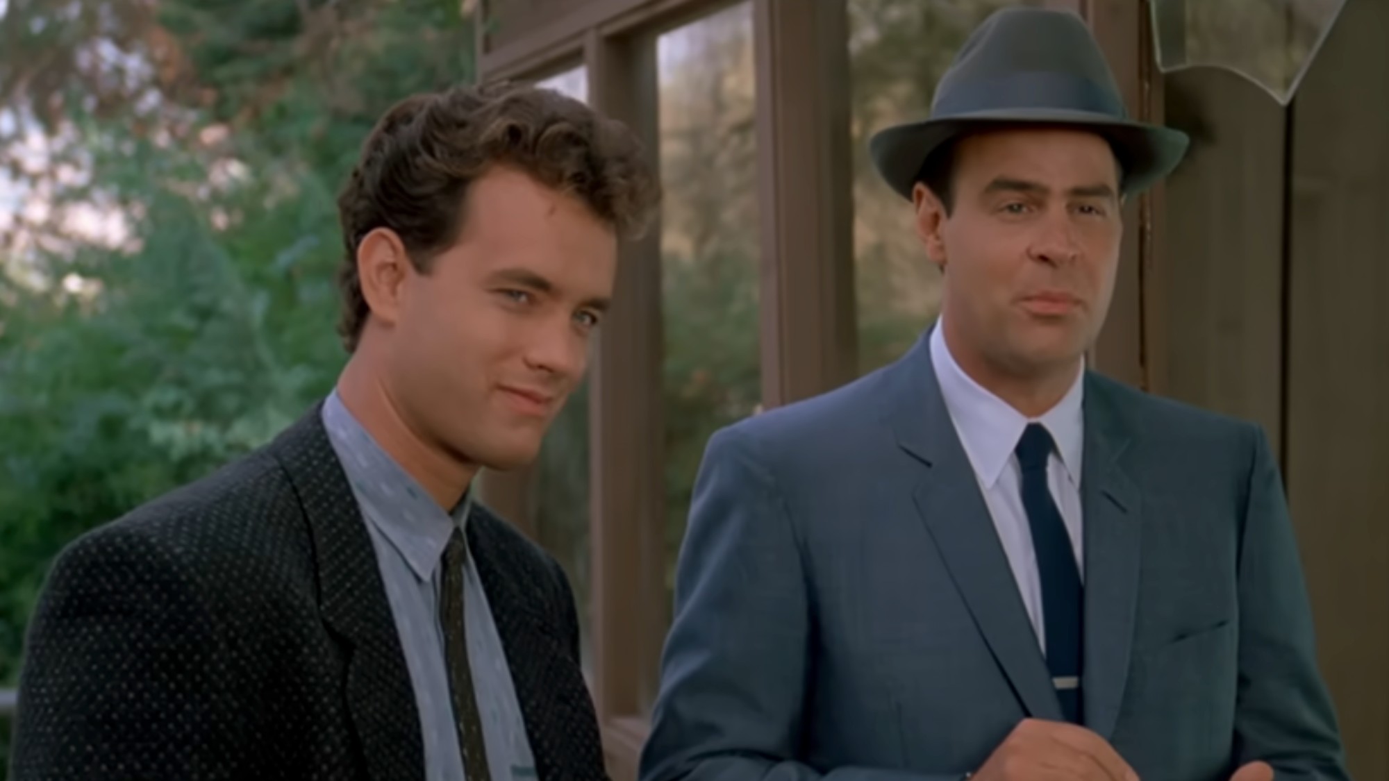 Tom Hanks smiling in a jacket and tie, standing next to Dan Aykroyd wearing a blue suit and fedora in Dragnet.