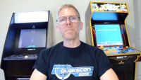 Man with glasses and a "QuakeCon 2008" shirt smiles in front of two arcade machines; one for Atari Asteroids, the other for Joust. Nostalgic gaming vibe.