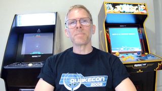 Man with glasses and a "QuakeCon 2008" shirt smiles in front of two arcade machines; one for Atari Asteroids, the other for Joust. Nostalgic gaming vibe.