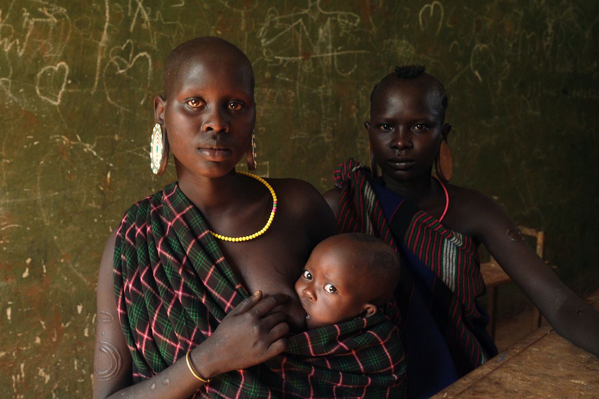 Two black women wearing traditional attire, with colorful jewelry, seated in front of a chalkboard, one holding a child wrapped in fabric