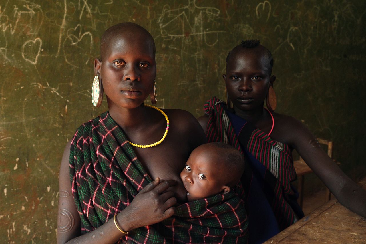 Two black women wearing traditional attire, with colorful jewelry, seated in front of a chalkboard, one holding a child wrapped in fabric