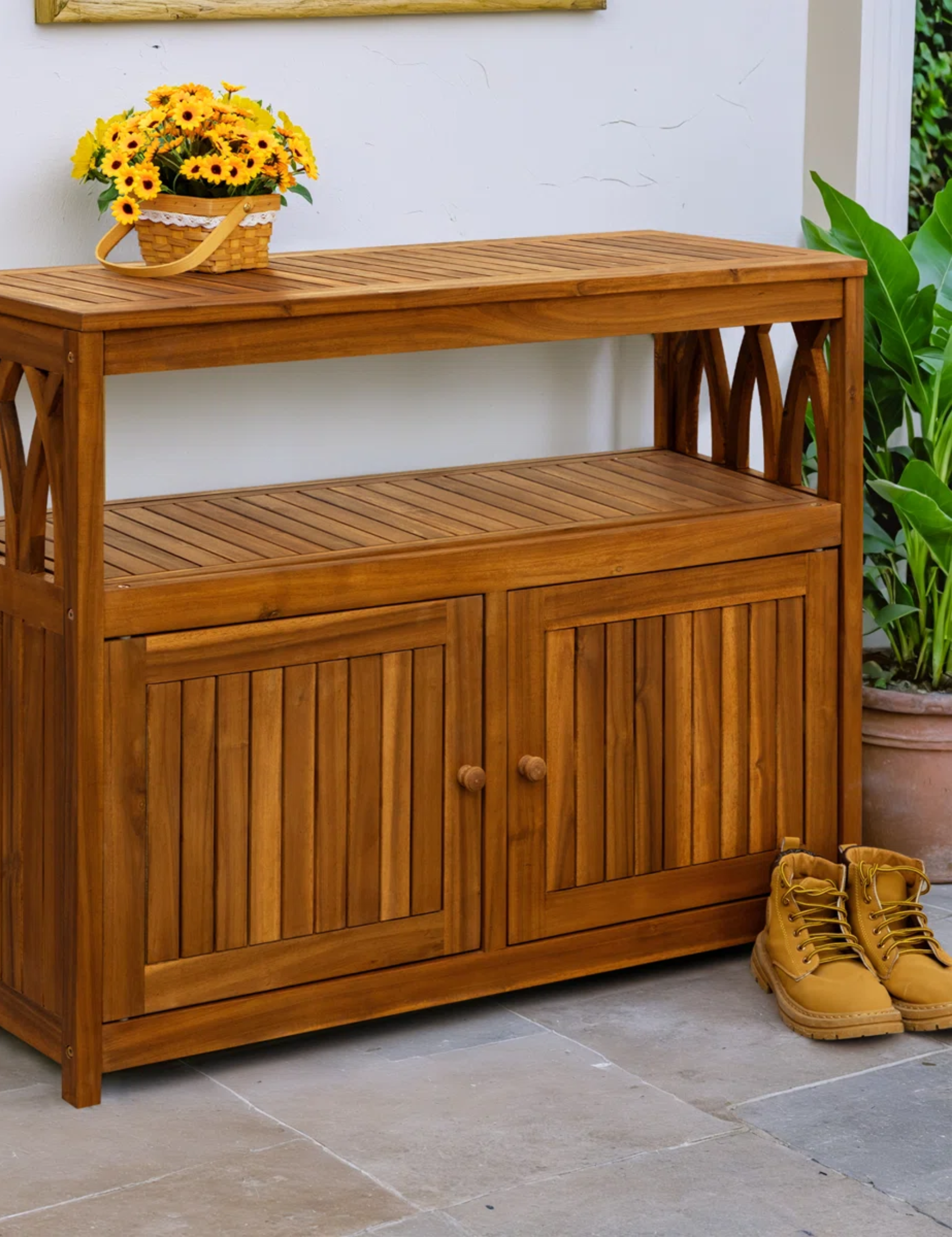 Wooden outdoor sideboard styled alongside a pair of work boots and a basket of sunflowers atop a stone-floored patio.