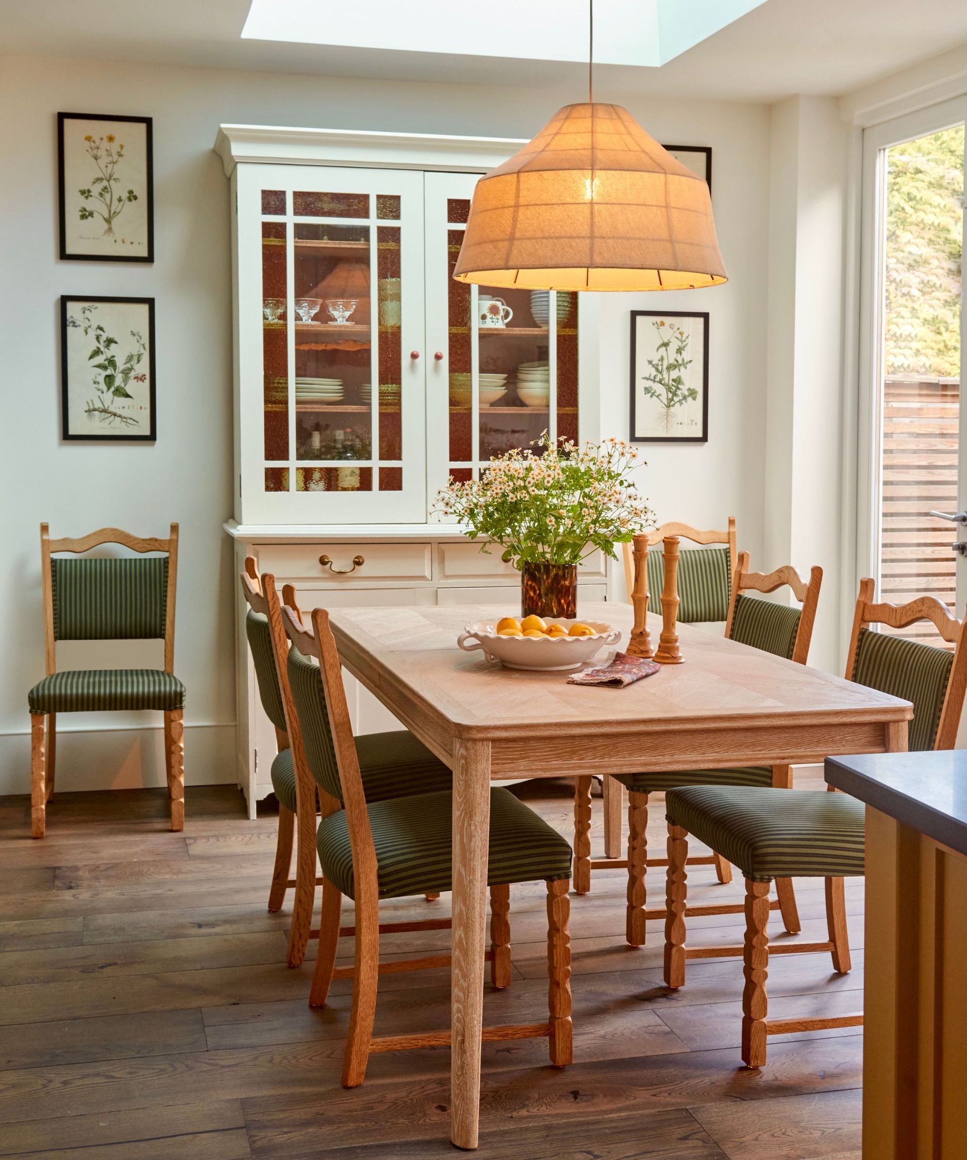 Dining room with vintage wood table and chairs, pendant light, wood cabinet with plates and glasses and vintage botanical prints