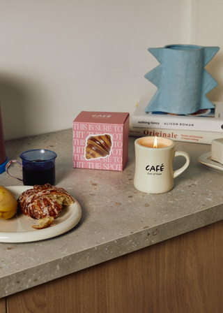 A terrazzo countertop with a couple of books, a textured blue vase, a mug of coffee, a glazed croissant on a plate, a butter dish and a mug candle alongside its boxed packaging.