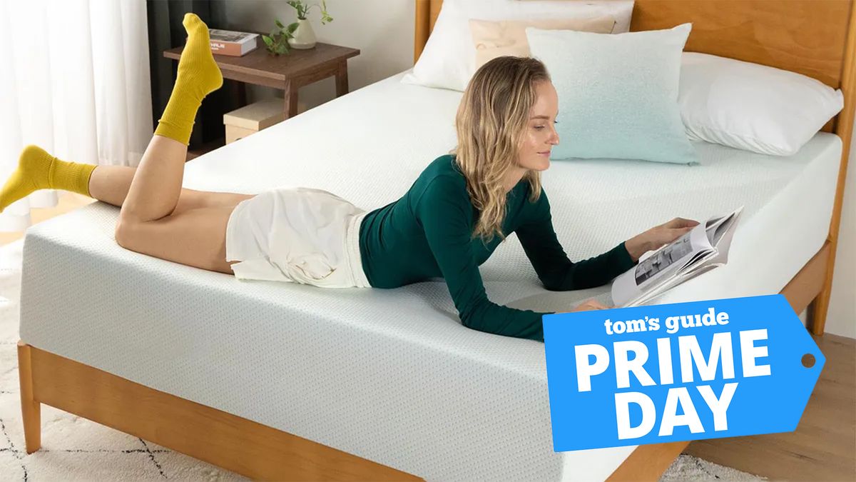 A woman in a green top and yellow socks lies on her stomach on the Zinus Green Tea Memory Foam Mattress while reading a book, with blue Prime Day deals badge in the bottom right hand corner