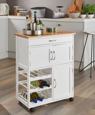 A compact white kitchen trolley with a natural bamboo top, featuring a built-in wine rack, a drawer, and a double-door storage cabinet. Small wheels provide easy mobility, and the top holds a tray with bottles of olive oil and seasoning.