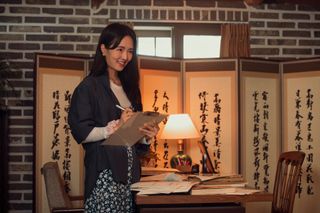 Hojo Shin as Jiwon standing by a desk and holding a clipboard in episode 205 of XO, Kitty.