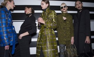 Five female models wearing looks from Bottega Veneta's collection. One model is wearing a blue check style coat with a belt. Next to her is a model wearing a dark patterned piece and neck scarf. The third model is wearing a dark green check style coat. The fourth model is wearing a dark green cardigan, grey patterned trousers and she is holding a bag. And the fifth model is wearing a black and grey patterned coat and she is also holding a bag