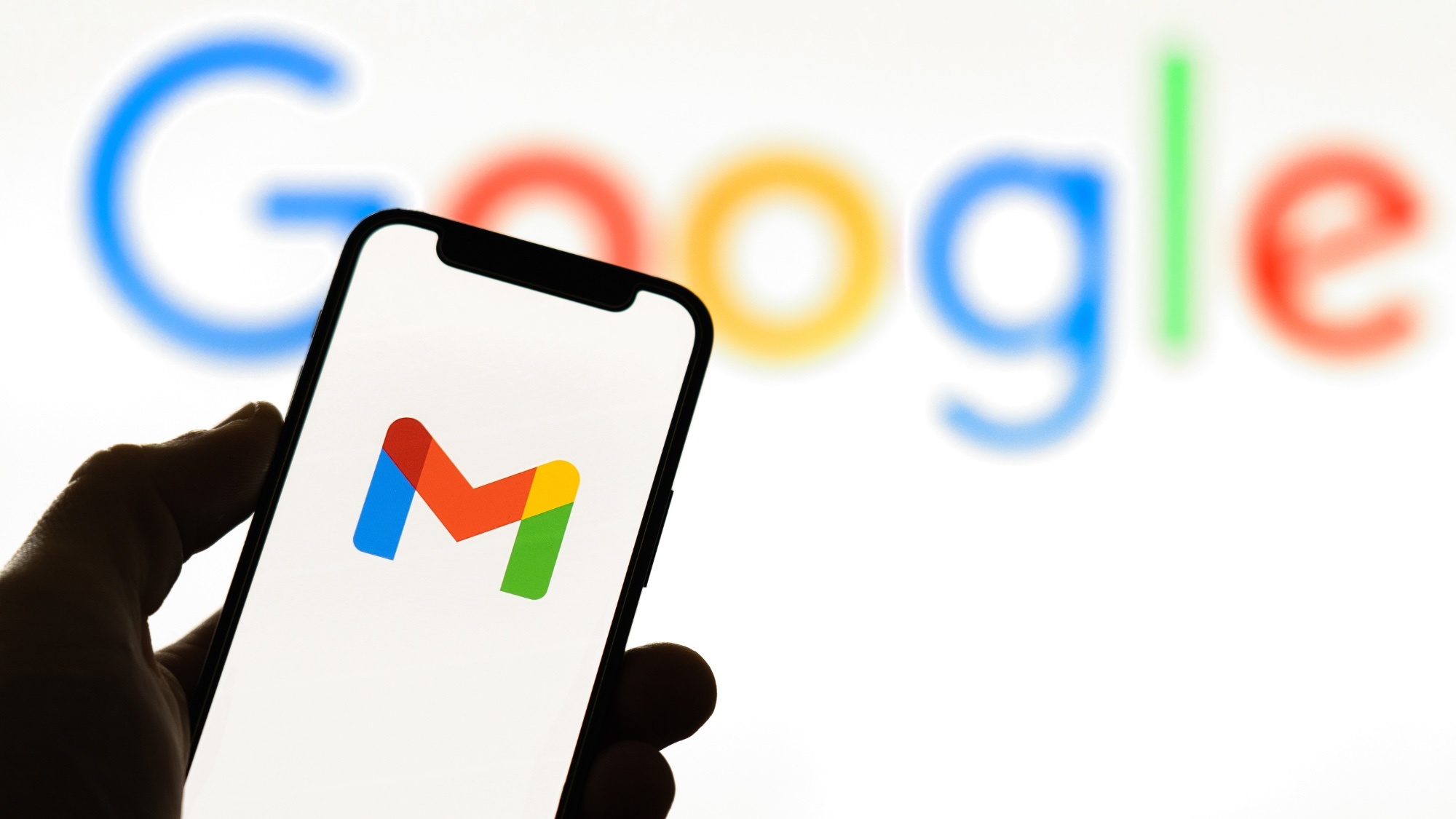 A smartphone with the Gmail logo on it, being held by a hand, in front of a Google background