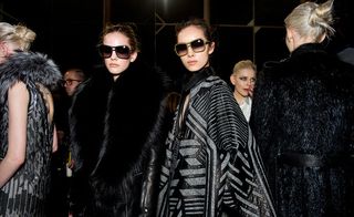 Four female models wearing looks from Roberto Cavalli's collection. One model is wearing a black and grey sleeveless piece with fur. Another model is wearing a black leather coat with fur. Next to her is another model wearing a black and grey cape style piece. And the fourth model is wearing a black belted fur jacket Two models are wearing sunglasses and there are other people in the background