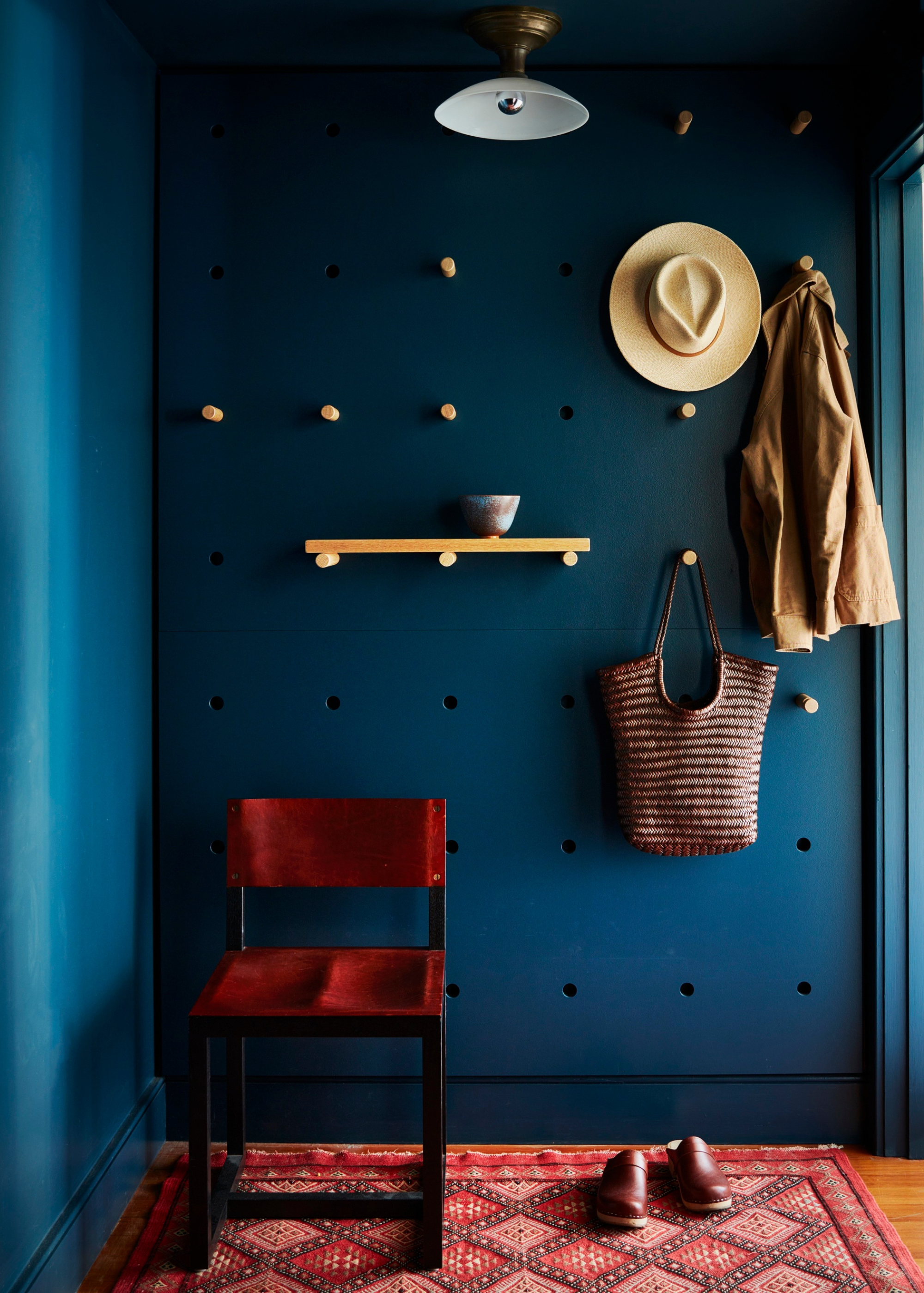 an entryway with a dark blue full-length wall pegboard with hooks, for a hat, coat, bag and mini shelf; there is a red chair in front of the wall on top of a red rug with shoes on top