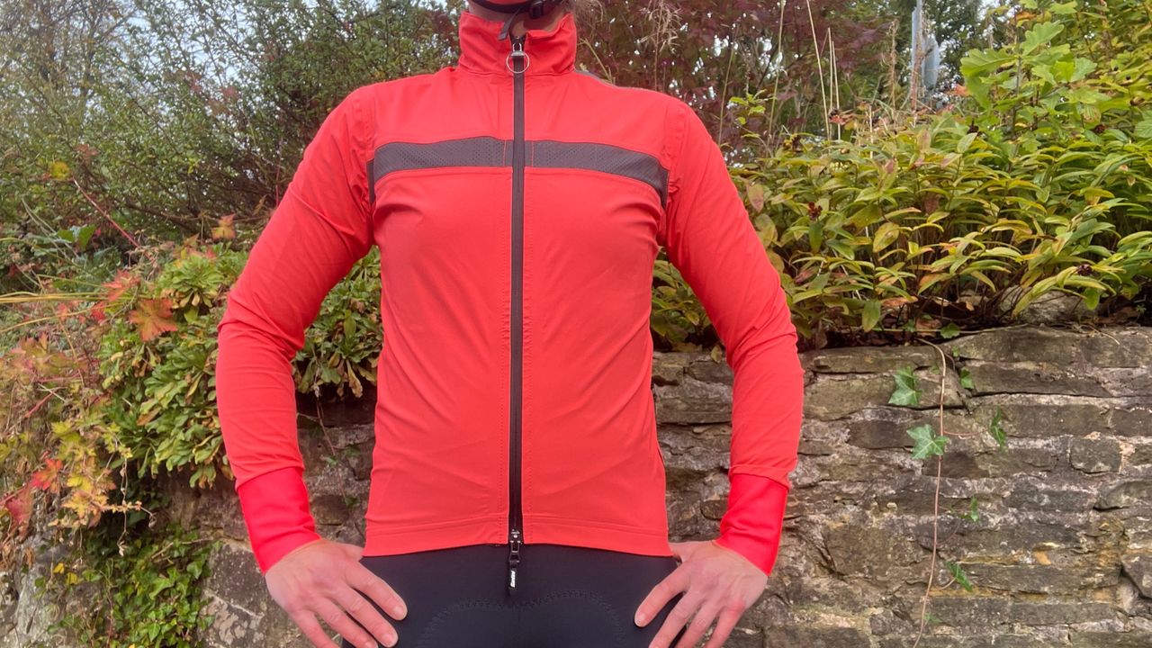 The Santini Guard Neos Rain Jacket in bright pink on a woman&#039;s torso standing in front of a wall and greenery 