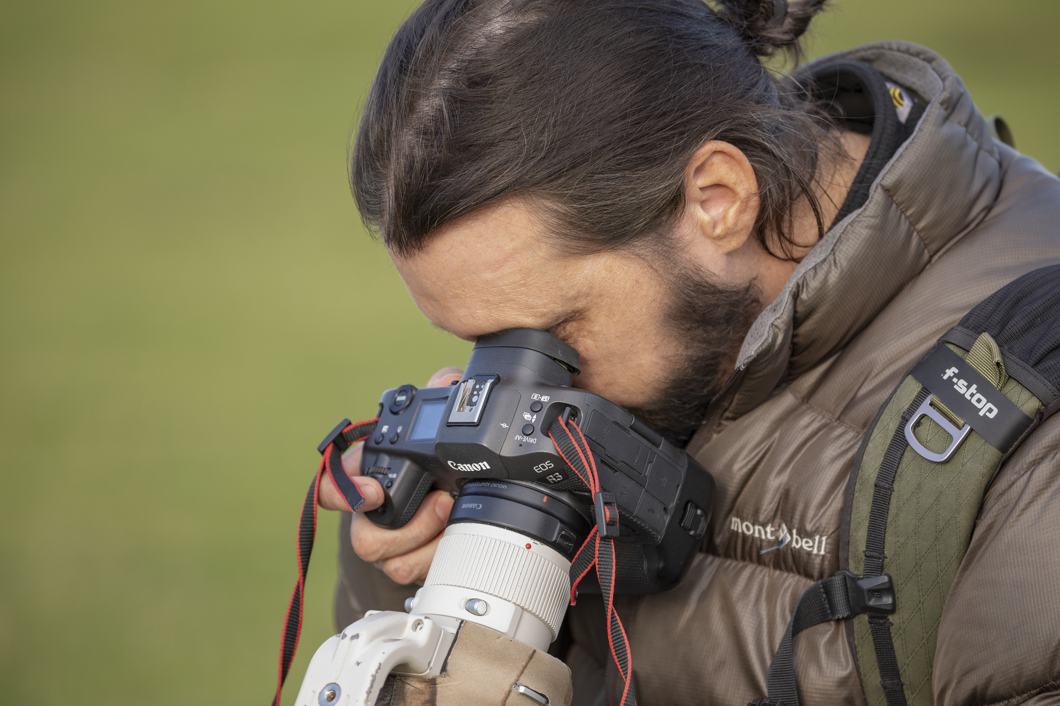 Photographer with dark hair in ponytail looks through EVF of a Canon EOS R3, greenery blurred behind