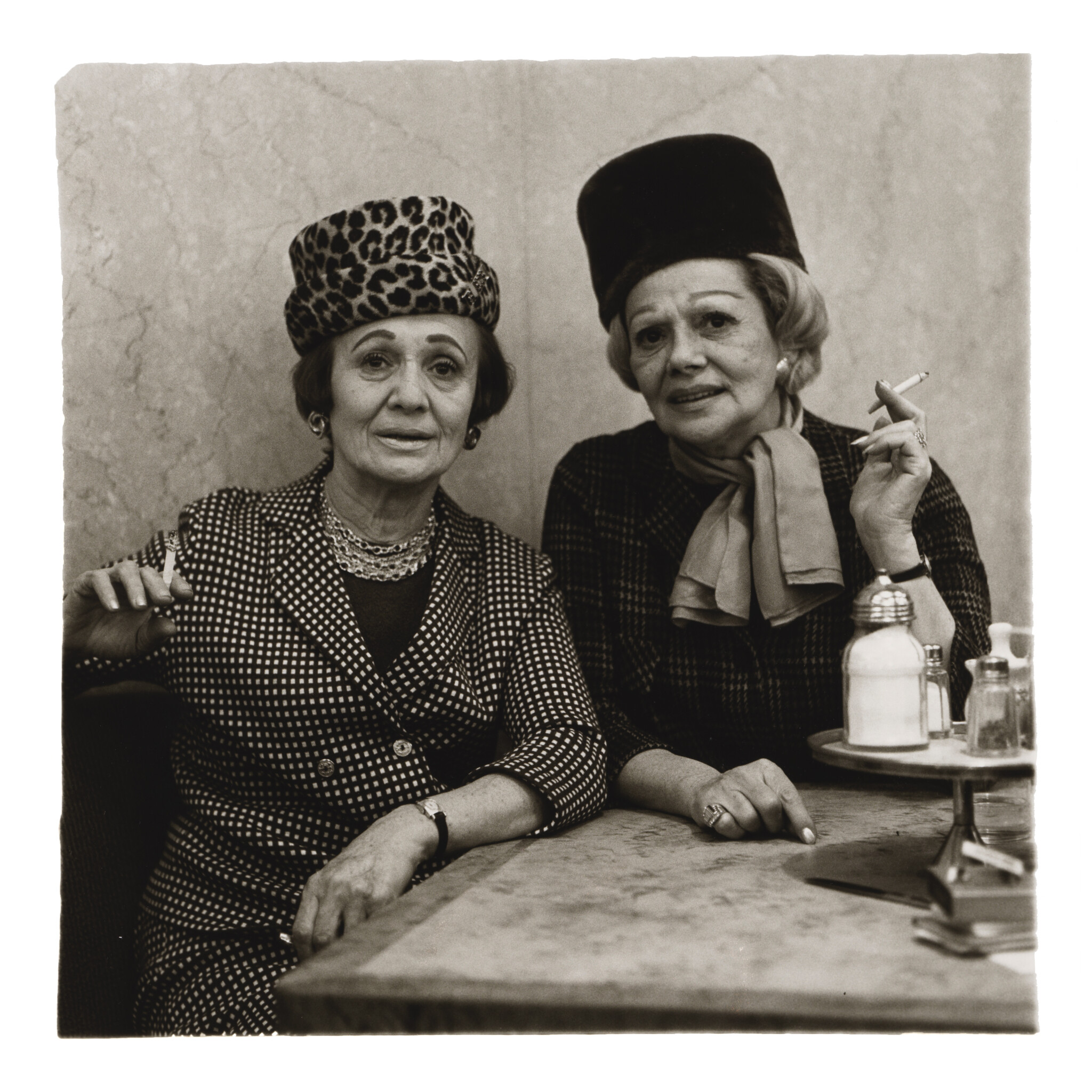 A black and white, close-up photograph features two women sitting at a cafe table wearing stylish hats and patterned suits while holding cigarettes.