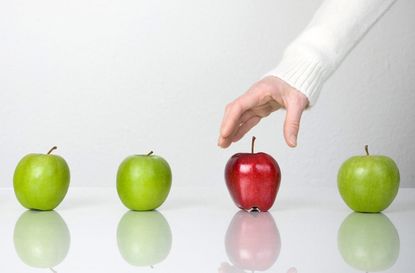 Out of four apples, three are green and one is red. A hand reaches for the red one.