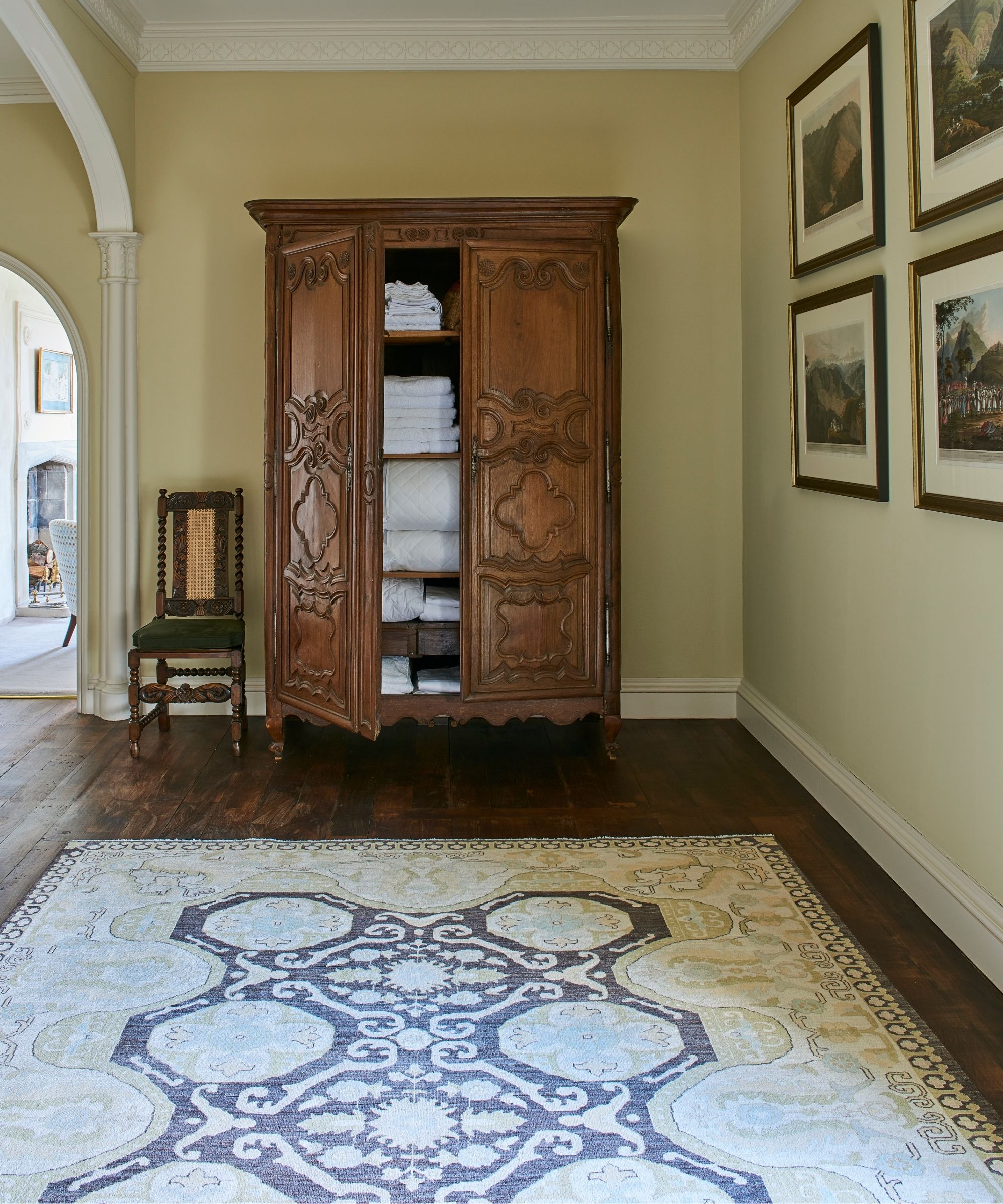 A large wooden linen closet pictured in a painted hallway with dark wood flooring, a large patterned rug, a wooden chair with dark green upholstering, and a landscape gallery wall.