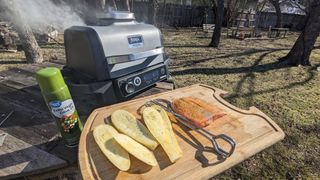 Salmon and squash on a wooden cutting board, next to a Ninja Woodfire Outdoor Grill, cooking outside