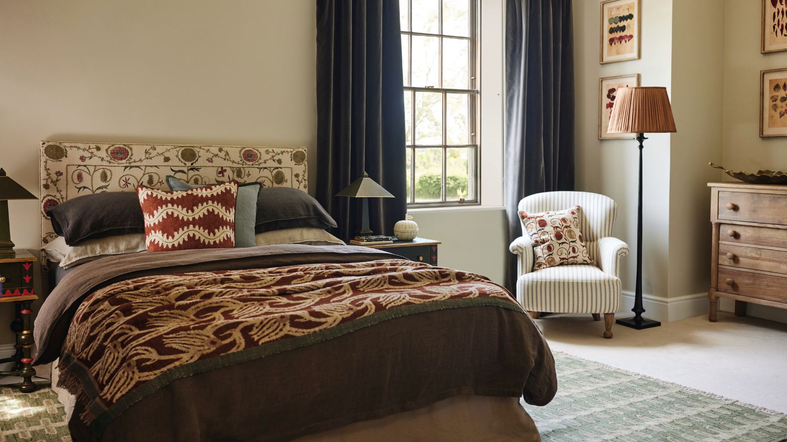 An autumnal bedroom with brown bedding, a patterned headboard, and a white chair beneath a lamp in the corner beside a window. 
