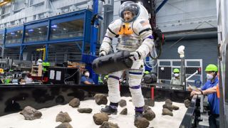 A NASA volunteer carries a 30-pound (14 kilograms) object while in a spacesuit connected to NASA’s Active Response Gravity Offload System (ARGOS).