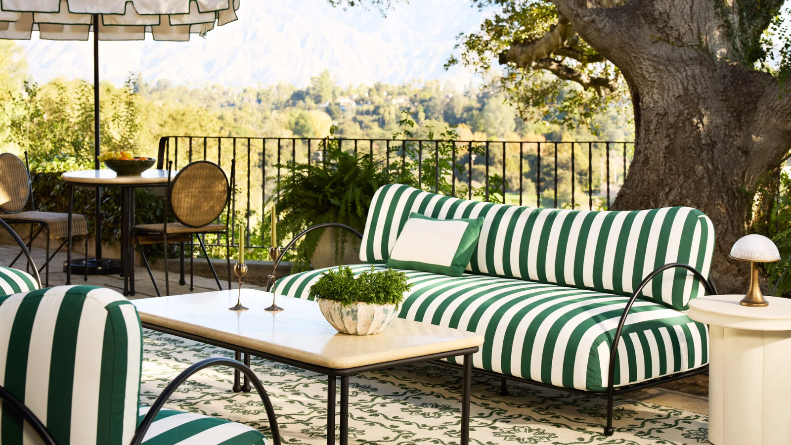a patio with a green and white striped couch, a travertine coffee table, and a bistro set with an umbrella all overlooking a woodland scene