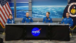 a woman and three men wearing blue coveralls sit at a table with the NASA logo on its front and flags to both sides.
