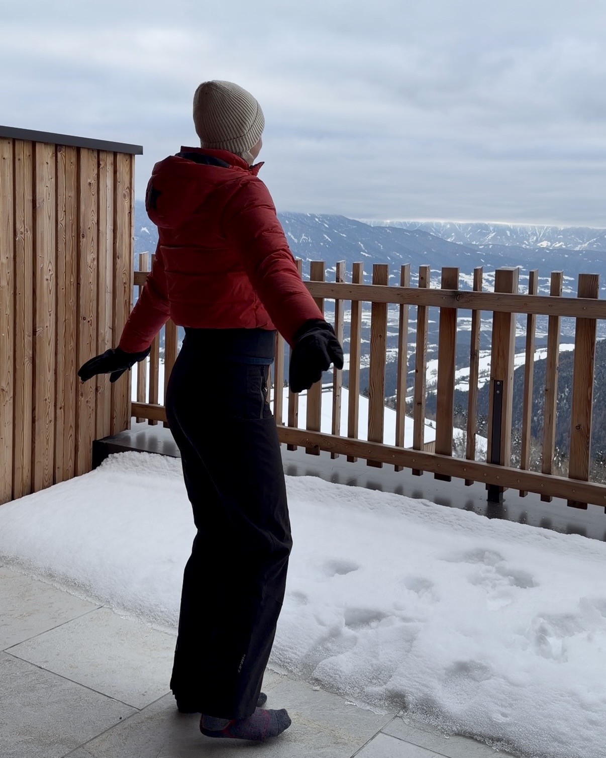 A woman in a red ski jacket stands on a snowy balcony