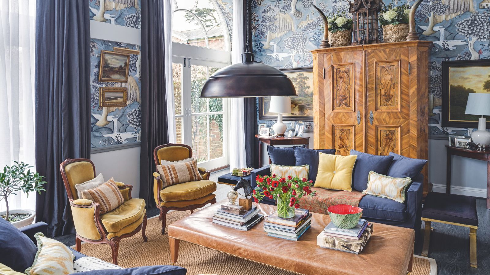 Blue and brown eclectic living room with blue patterned wallpaper. In the centre of the room is a low, large leather ottoman with stacks of books and flowers. There is a blue sofa and two matching velvet upholstered chairs. Behind is a wooden cupboard and large french doors filling the room with natural light.
