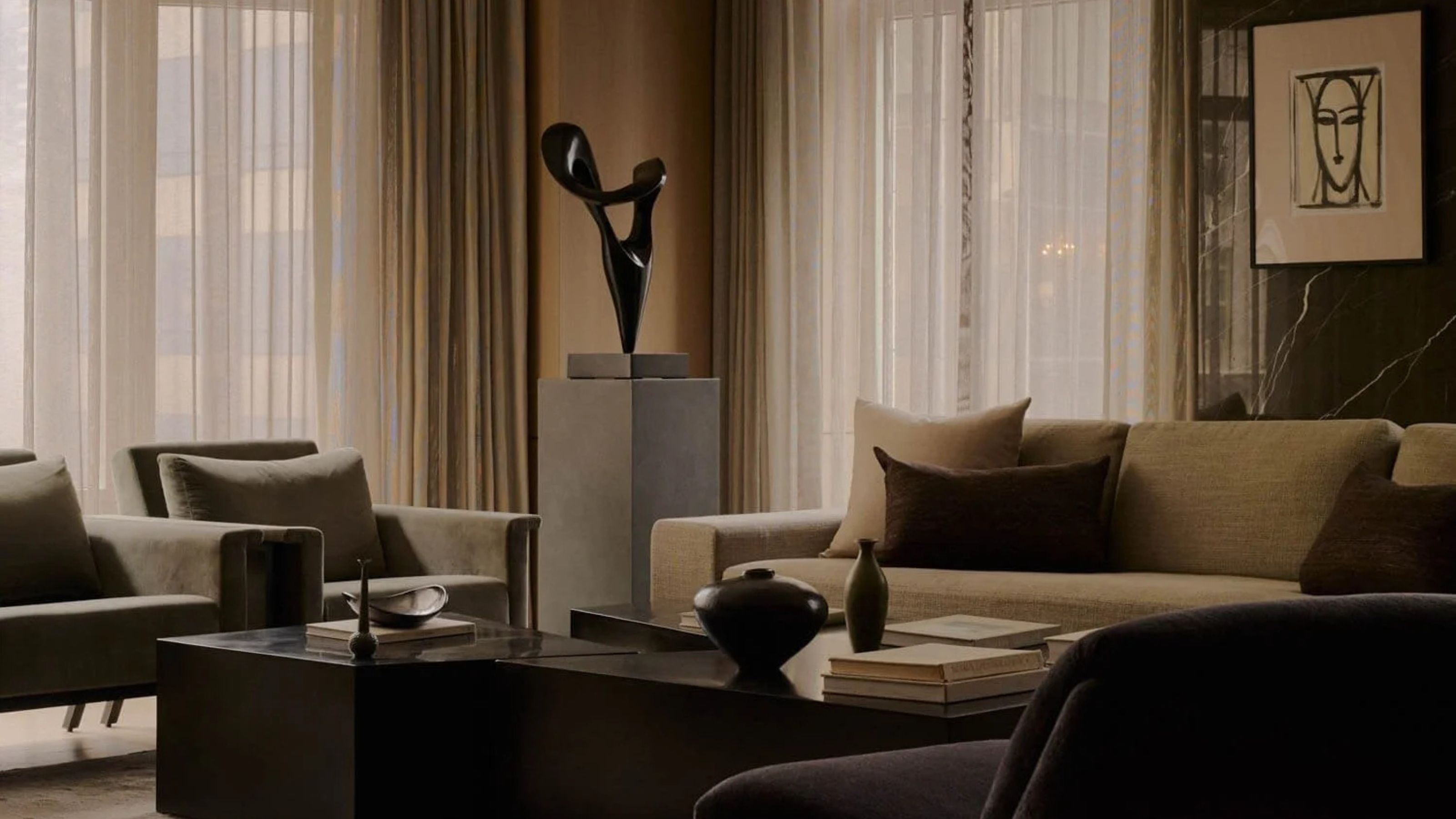 A living room with a sheer curtains that have been closed. In the living room with a large sofa, accent chairs, a coffee table as well as a large piece of artwork/sculpture beside the sofa sofa and accent chair.