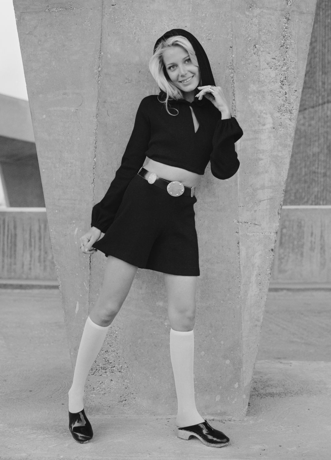 A young female model wears black French shorts and cropped bolero style top with hood, knee length socks and clogs in front of a concrete flyover in London on 22nd October 1970.