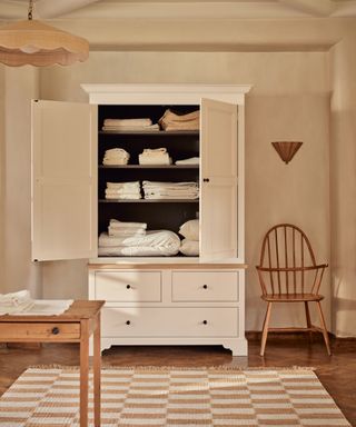 freestanding linen cupboard with bedding and pillowcase, stone colored walls, table, chair, rug, pendant