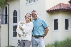 Portrait of an affluent mature couple standing in front of their home, a grand mansion. The woman is holding a cute pampered little dog in her arms while her husband stands beside her with his arm around her. They are smiling at the camera.