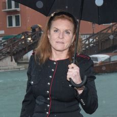 Sarah Ferguson wears a black jacket with metal buttons and red piping and holds a black umbrella while standing in the rain in Venice
