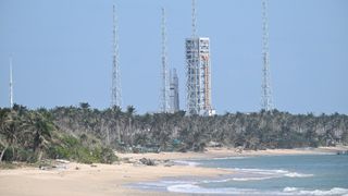 a tropical beach lined with palm trees in front of a rocket upright on a launch pad in the background