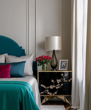 A black nightstand with a black and white lamp and flowers on next to a bed with teal oval blue and silver and teal silk bedsheets