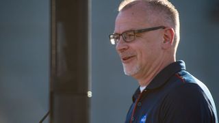 a man wearing a dark nasa logo polo shirt and glasses smiles with a greying goatee.