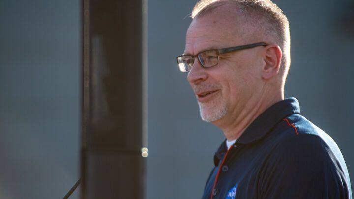 a man wearing a dark nasa logo polo shirt and glasses smiles with a greying goatee.